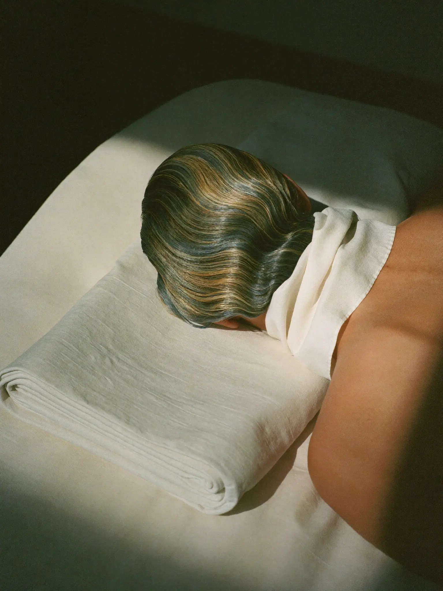 The crown of a client's head at rest on folded linen, face turned away, waved hair catching warm light